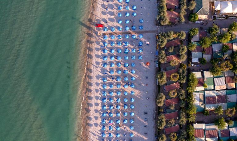 Spiaggia con ombrelloni blu e case vicine, vista dall'alto.