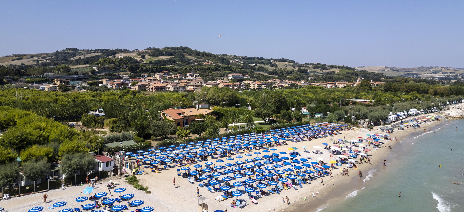 Plage bondée avec parasols bleus et mer, collines en arrière-plan.