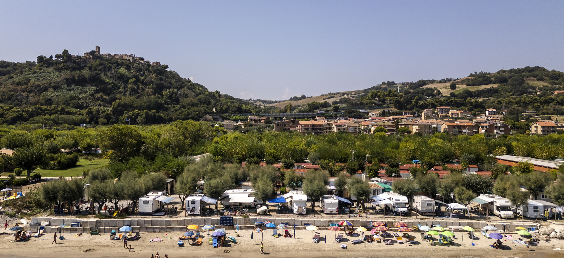 Plage avec parasols et camping-cars, collines et maisons en arrière-plan.